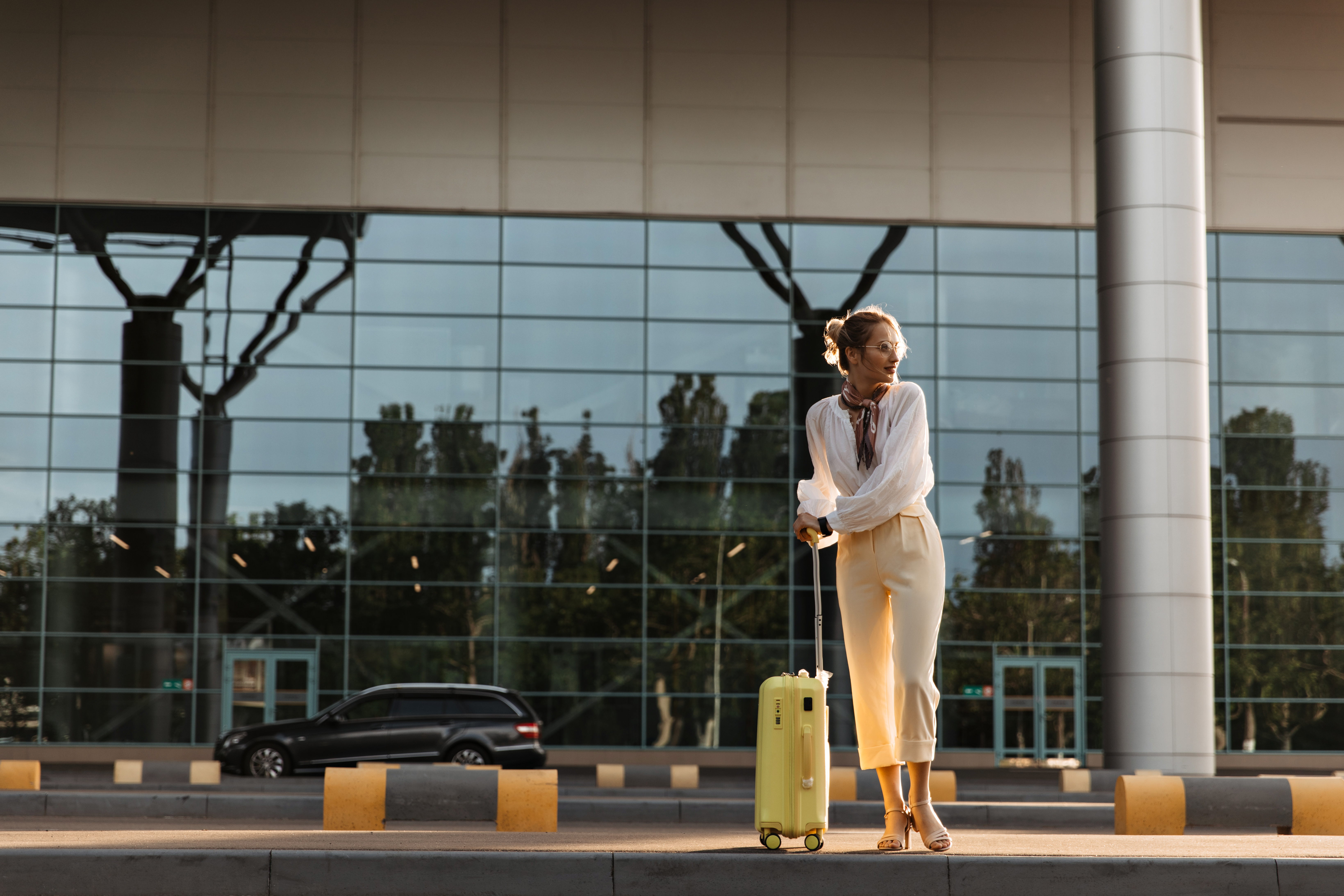Charming blonde woman in white blouse, beige pants and eyeglasses moves near airport. Pretty girl in stylish outfit holds yellow suitcase.